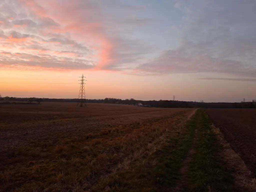 Twilight over a vast field with a dirt path, featuring a distant power line tower against a colorful sky filled with pink and blue clouds. Peaceful atmosphere.