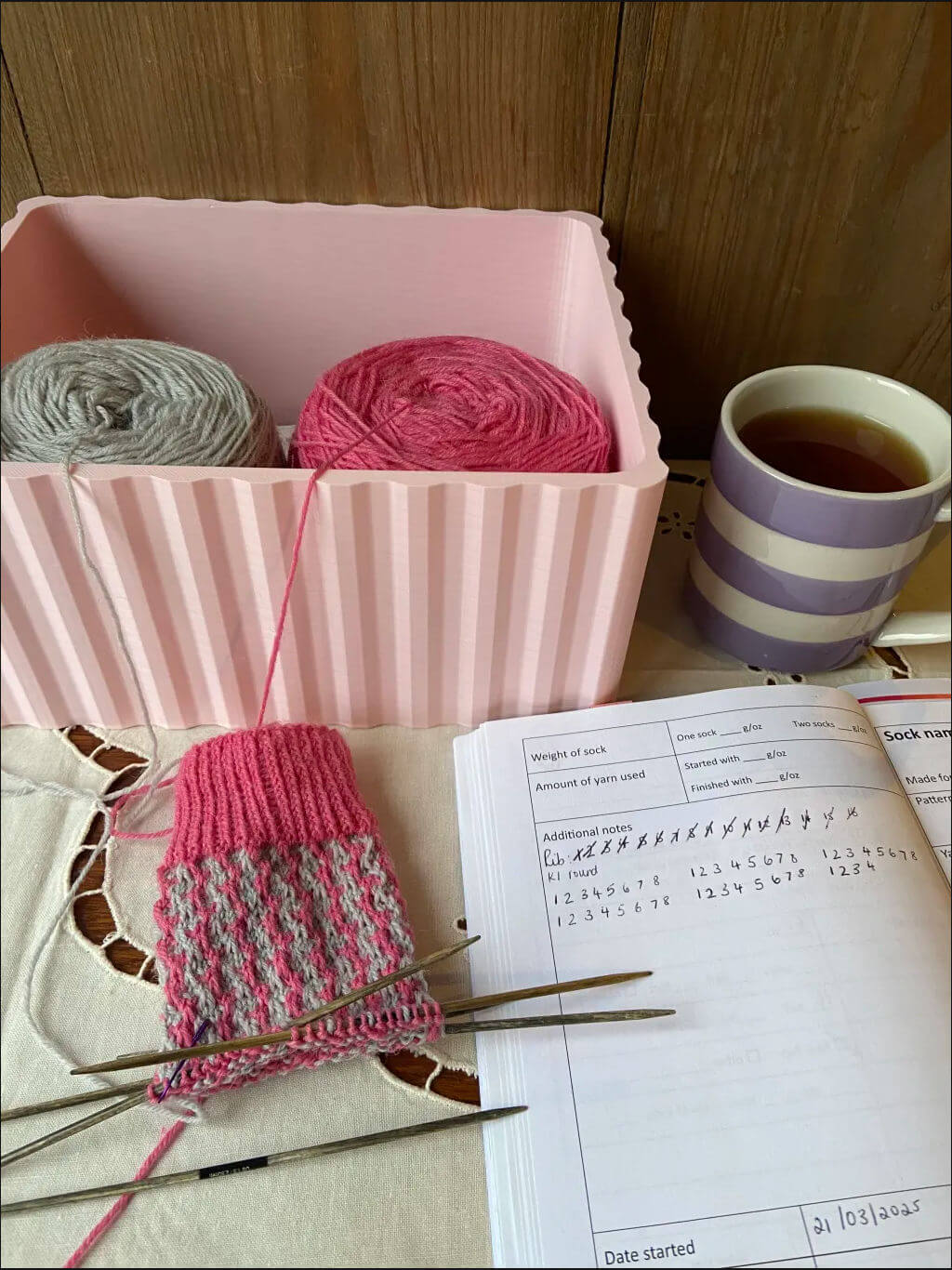 A cozy knitting scene shows pink and gray yarn in a pink box, a half-knitted sock on needles, knitting instructions, and a striped mug of tea.