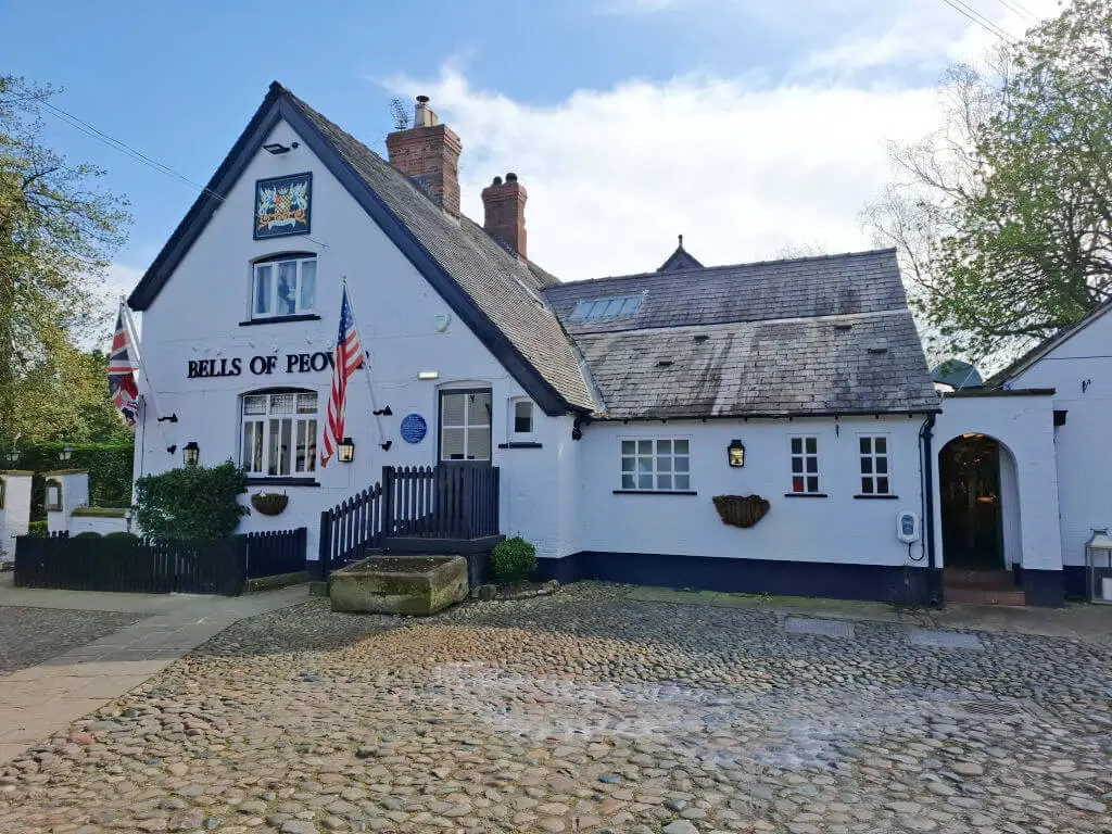Charming white-painted pub with flags outside, labeled "Bells of Peover." Cobblestone path leads to black picket fence. Bright, inviting atmosphere.