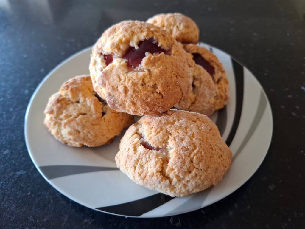 A plate with five round jam buns, some with visible caramelized jam, placed on a dark counter. The jam buns appear golden-brown, evoking a homemade, cozy feel.