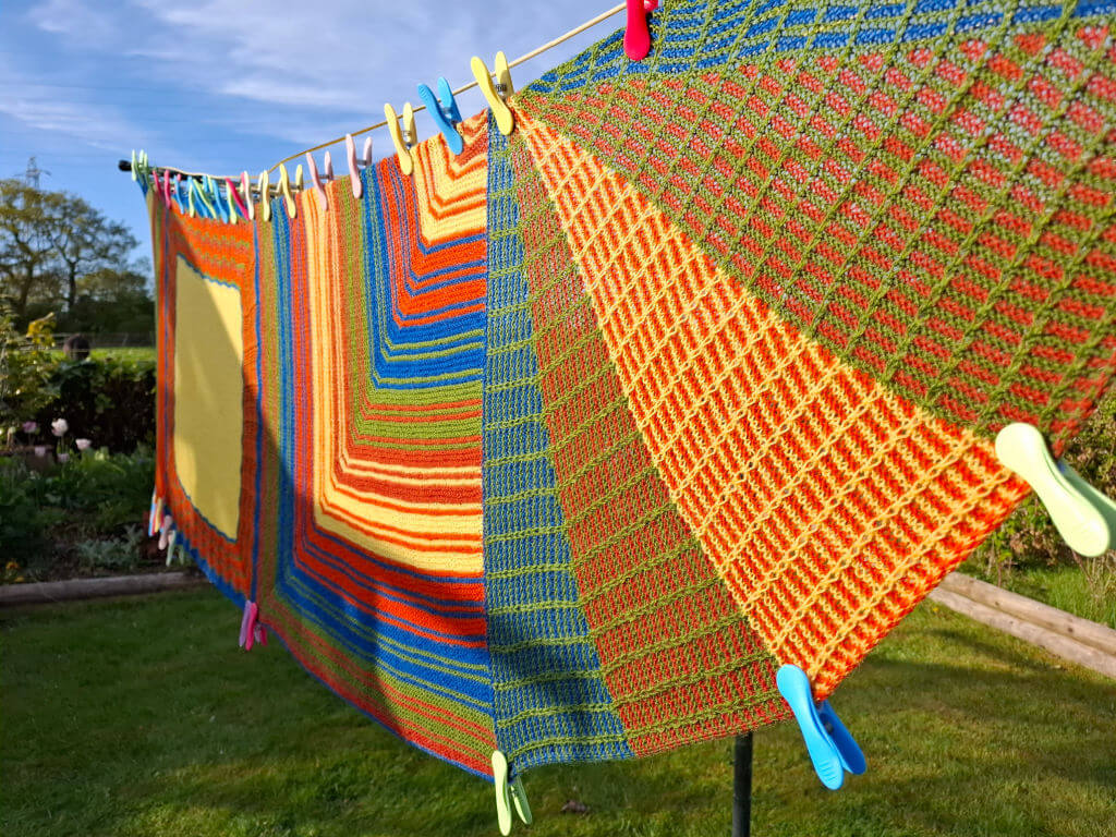 Brightly coloured knitted shawl with geometric patterns hangs on a clothesline with multicoloured pegs under a clear blue sky in a sunny garden.