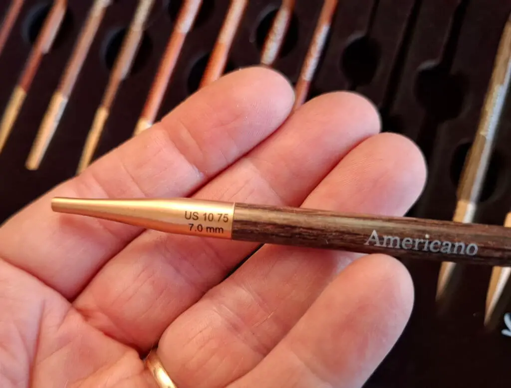 Close-up of a hand holding a knitting needle labeled "Americano," size US 10.75, 7.0 mm. Multiple needles are blurred in the background | Winwick Mum KnitPro Knit & Sip Set Review