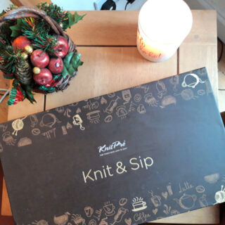 A "Knit & Sip" kit box lies on a wooden table, adorned with playful knitting and coffee-themed illustrations. Beside it, a festive arrangement with red apples and pinecones, and a glowing candle create a cozy, holiday ambiance.