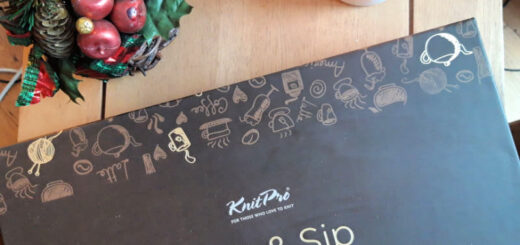 A "Knit & Sip" kit box lies on a wooden table, adorned with playful knitting and coffee-themed illustrations. Beside it, a festive arrangement with red apples and pinecones, and a glowing candle create a cozy, holiday ambiance.