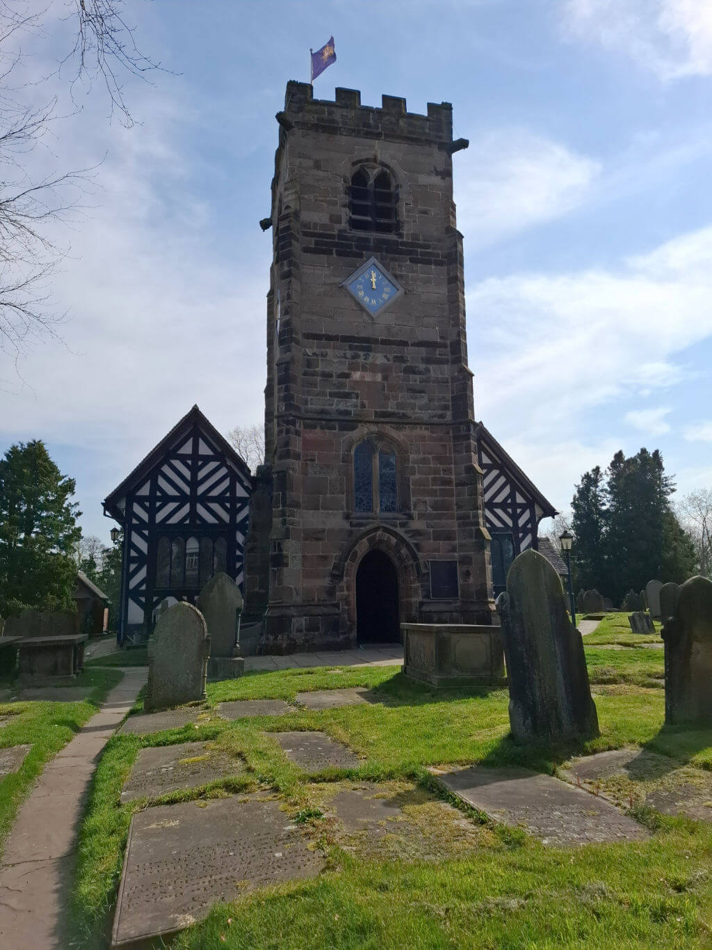 Historic stone church tower with clock and flag, surrounded by old gravestones on green grass, under a partly cloudy blue sky, evoking a serene atmosphere.