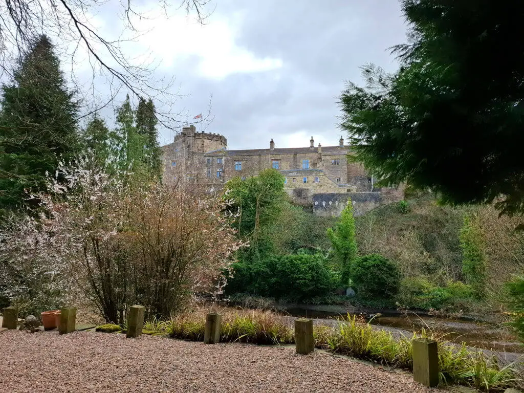 The majestic Skipton castle stands atop a hill, surrounded by lush greenery and blossoming trees. The overcast sky adds a calm, historical ambiance.