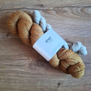 Two skeins of yarn, one golden brown and the other light gray, lie on a wooden surface. A label reads "ginger pink," conveying a rustic, cozy vibe.