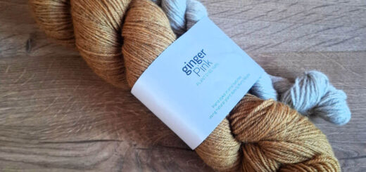 Two skeins of yarn, one golden brown and the other light gray, lie on a wooden surface. A label reads "ginger pink," conveying a rustic, cozy vibe.