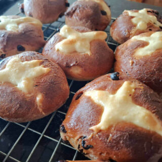 Freshly baked hot cross buns on a cooling rack, featuring golden brown tops marked with white flour crosses and scattered raisins, evoke warmth.