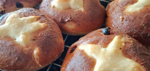 Freshly baked hot cross buns on a cooling rack, featuring golden brown tops marked with white flour crosses and scattered raisins, evoke warmth.