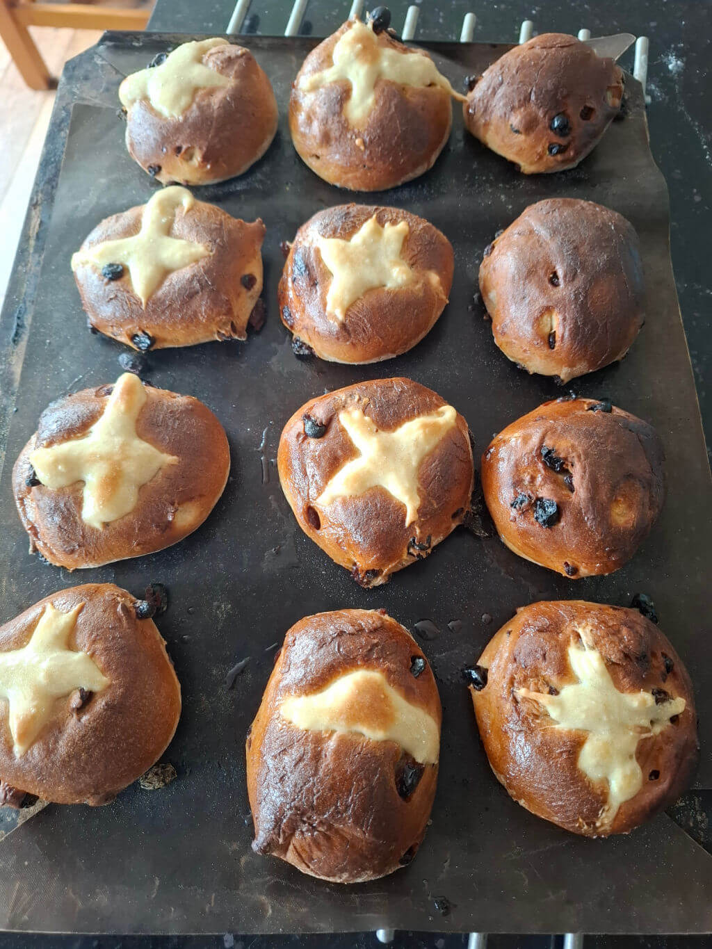 Twelve golden-brown hot cross buns with raisin specks on a baking tray, each adorned with a white cross, convey a fresh-baked warmth.
