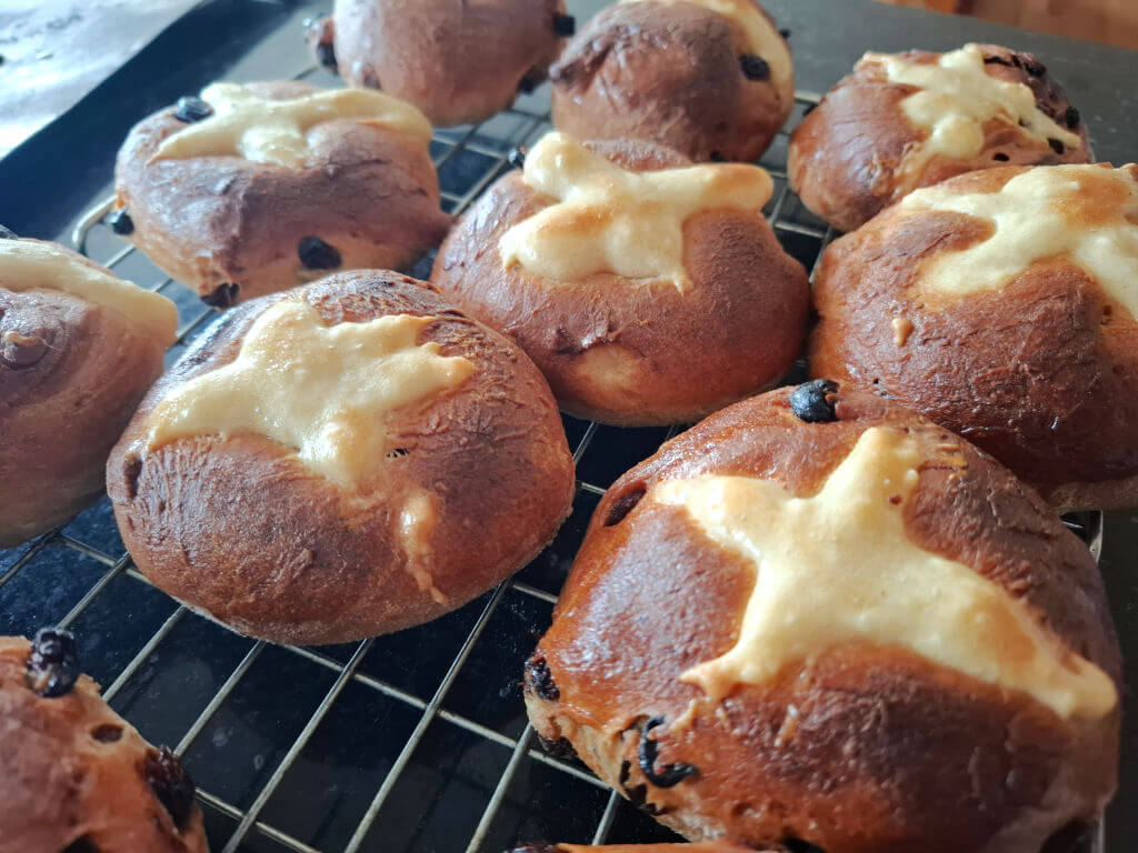 Freshly baked hot cross buns on a cooling rack, featuring golden brown tops marked with white flour crosses and scattered raisins, evoke warmth.