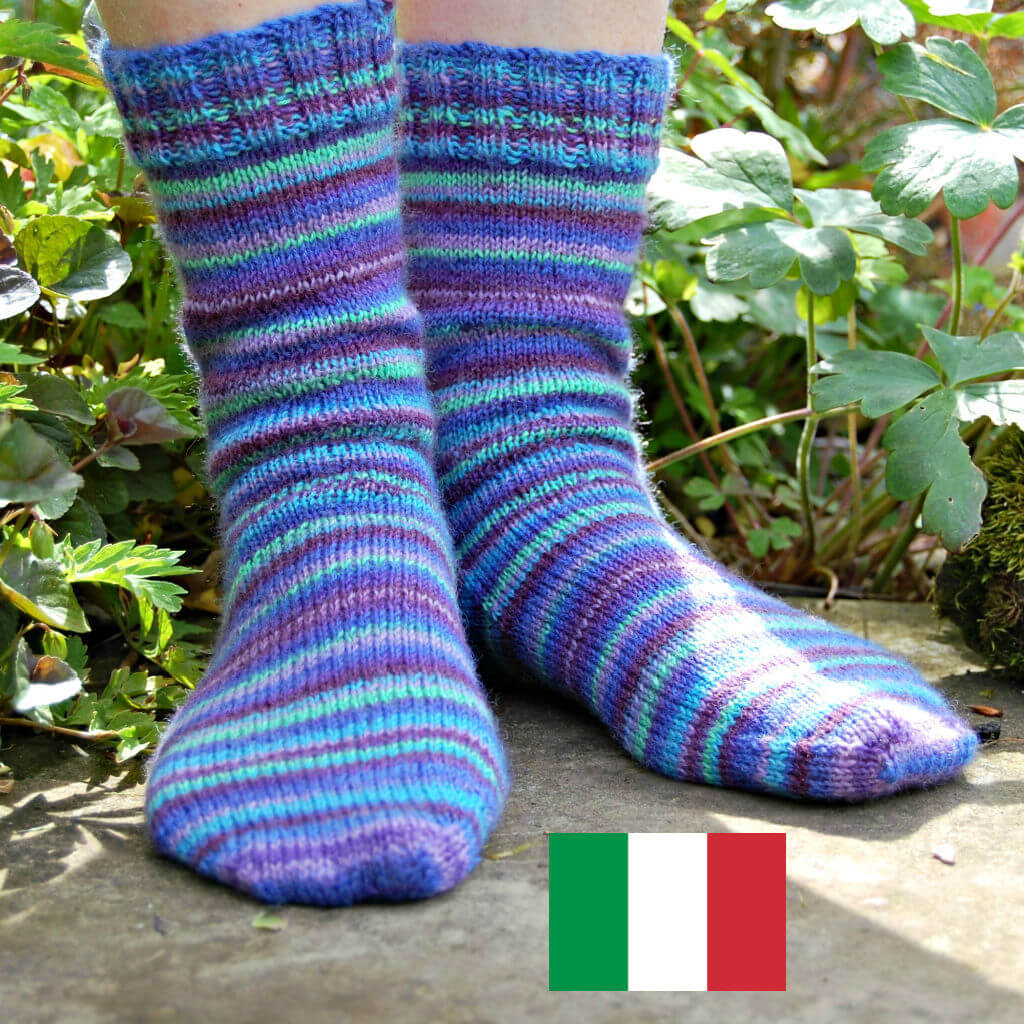 Primo piano di piedi che indossano calzini lavorati a maglia con strisce blu, verdi e viola, appoggiati su una superficie di pietra circondata da verde. Winwick Mum | Calzini Basic a 4 capi | Calzini per principianti | Schemi gratuiti per calzini