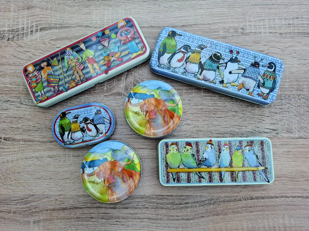 Colourful tin boxes with whimsical animal illustrations are displayed on a wooden surface. Designs include penguins, birds, and a highland cow, creating a playful and artistic tone by artist Emma Ball.