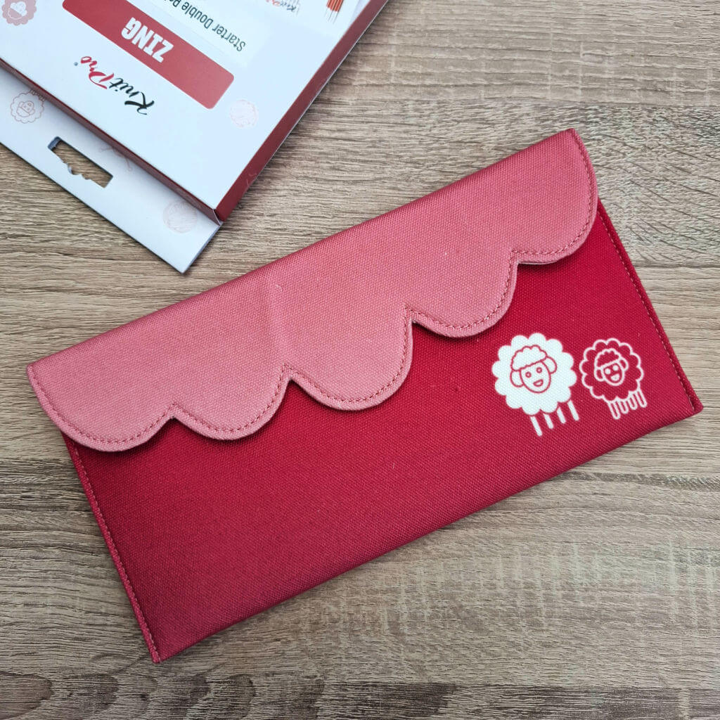 A pink and red scalloped fabric pencil case with two cute sheep designs lies on a wooden surface. Nearby is knitting packaging. The tone is playful. KnitPro Zing DPN review
