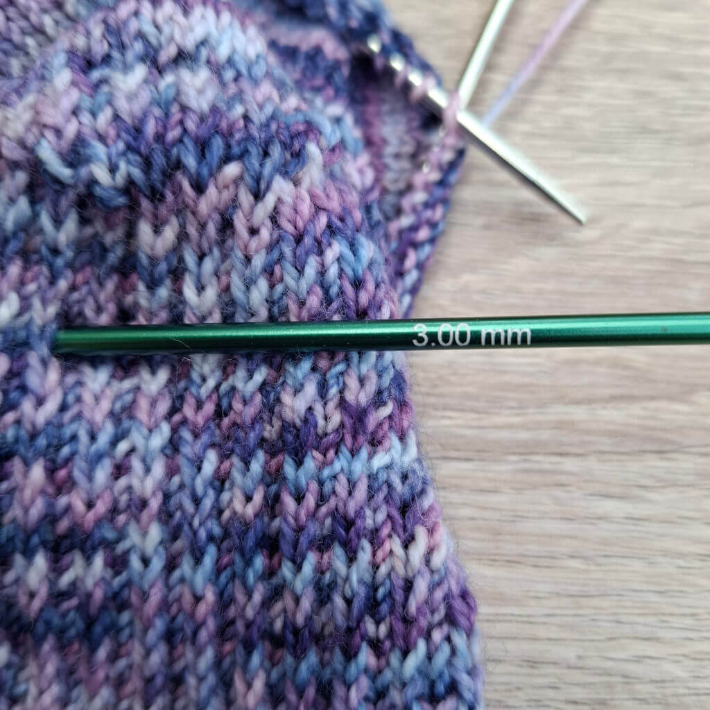 A knitting needle labeled "3.00 mm" rests on purple and blue knitted fabric with a woven texture, on a light wooden surface in soft focus.
