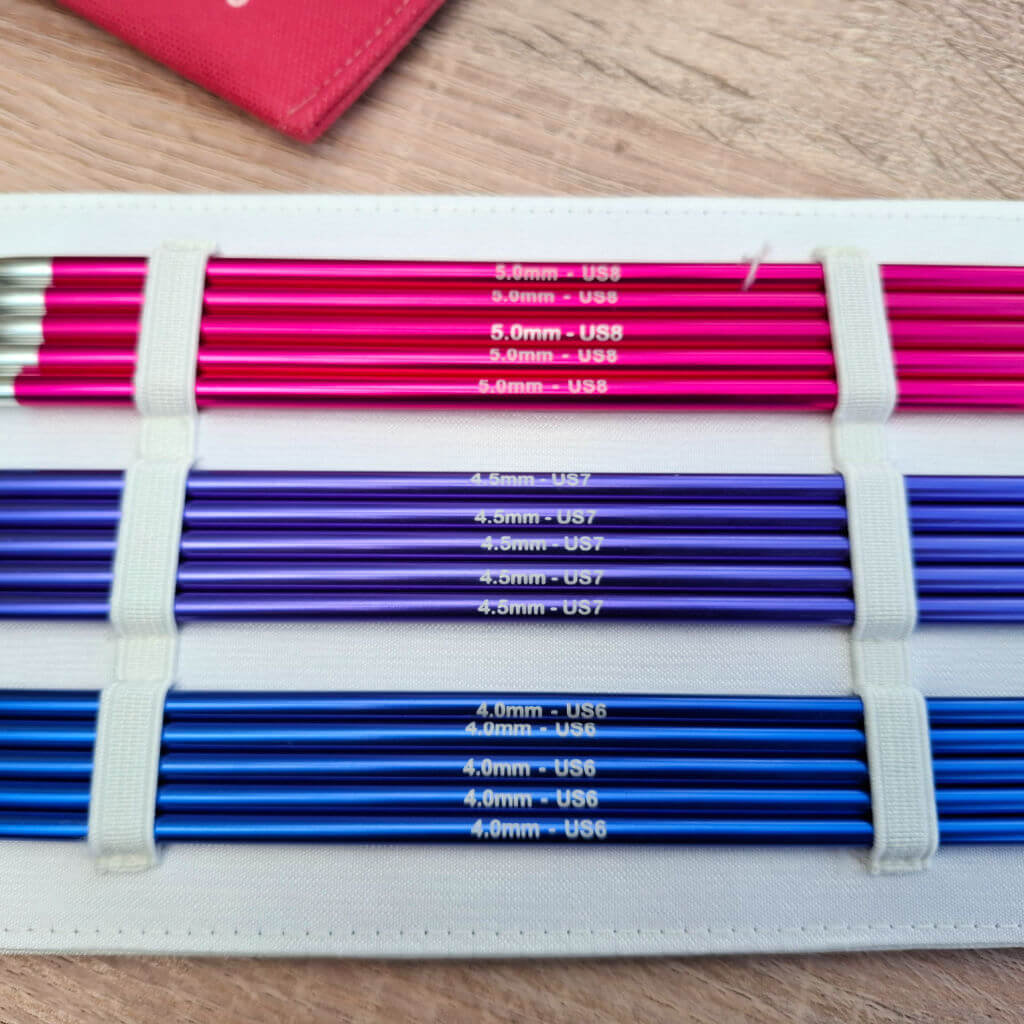 A set of colourful knitting needles organised by size in a white case. The top row is pink, the middle is purple, and the bottom is blue.