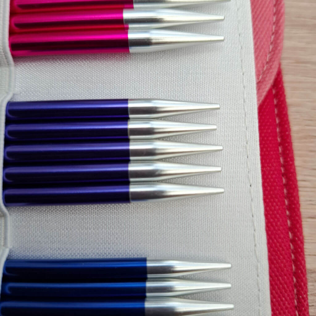 Close-up of nine metallic knitting needle tips aligned in a case. Handles are red, purple, and blue. The background is a textured white fabric. Red and pink knitting needle cases with sheep designs, red knitting needles, colourful yarn balls, and a wooden background. Cozy and creative vibe.