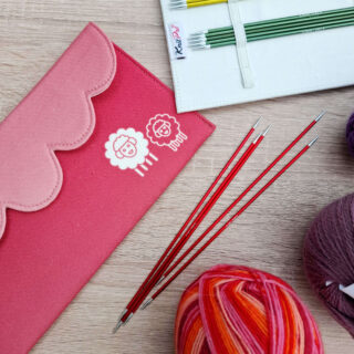 Red and pink knitting needle cases with sheep designs, red knitting needles, colourful yarn balls, and a wooden background. Cozy and creative vibe. KnitPro Zing DPN review