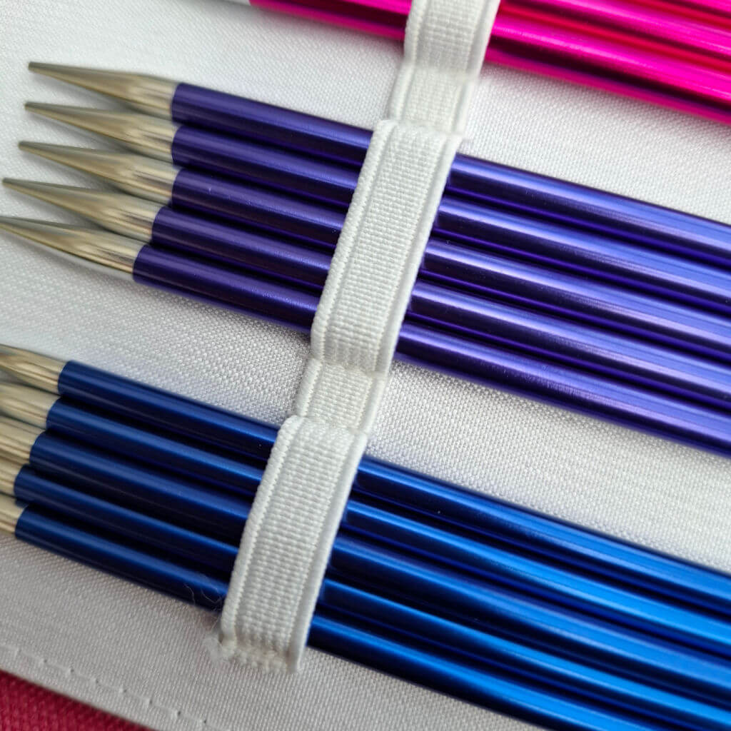 Close-up of colourful knitting needles in shades of blue, purple, and pink, neatly secured by white elastic bands on a fabric background.
