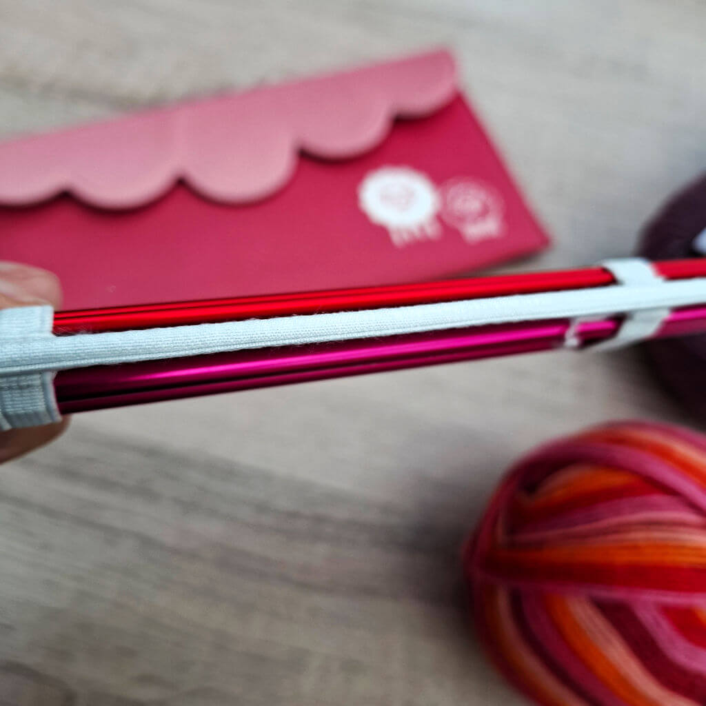 Close-up of red and pink knitting needles bound together with white elastic. A pink scalloped pouch and colourful yarn balls are in the background. KnitPro Zing DPN review