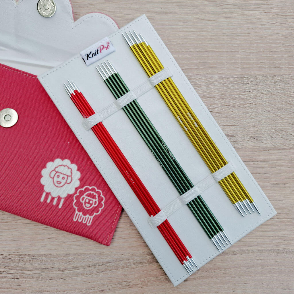 A knitting needle set with red, green, and yellow metal needles in a white holder. Next to it, a pink fabric case with sheep designs on a wooden surface. KnitPro Zing DPN review