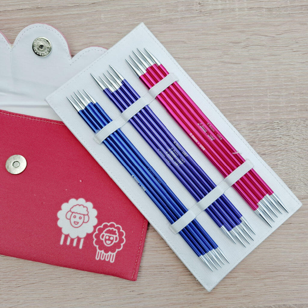 Knitting needle set with blue and pink metal needles in a white holder rests on a table. A partially open red case features cute sheep illustrations. KnitPro Zing DPN review