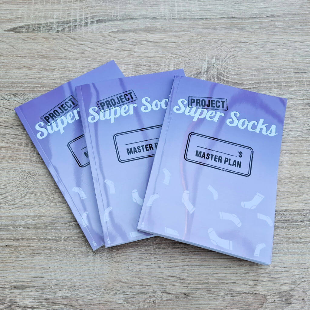 Three purple booklets titled "Project Super Socks: Master Plan" are fanned on a wooden surface. The design features scattered white sock illustrations.
