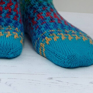 Close-up of colourful hand-knitted socks with a bright blue base and intricate red and yellow patterns. The socks rest on a white wooden surface.