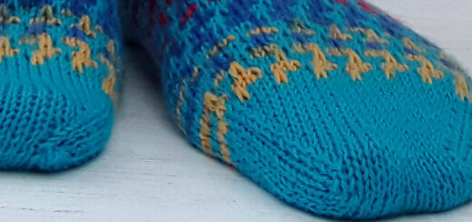 Close-up of colourful hand-knitted socks with a bright blue base and intricate red and yellow patterns. The socks rest on a white wooden surface.