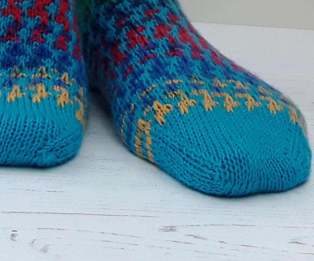 Close-up of colorful hand-knitted socks with a bright blue base and intricate red and yellow patterns. The socks rest on a white wooden surface.