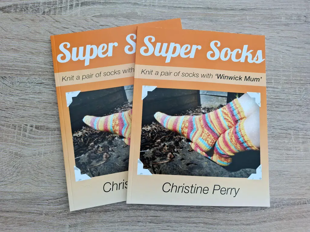 Cover of the book "Super Socks" by Christine Perry, featuring colorful knitted socks on a wooden floor. The tone is warm and inviting.