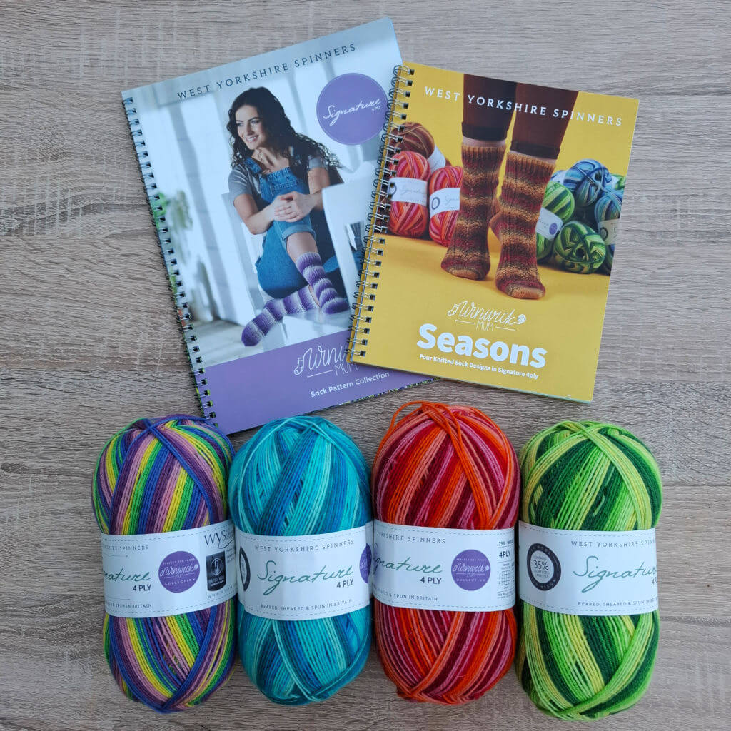 Four balls of colourful yarn, arranged in a row, sit below two pattern booklets: one featuring striped socks and a person, the other with vibrant sock designs.