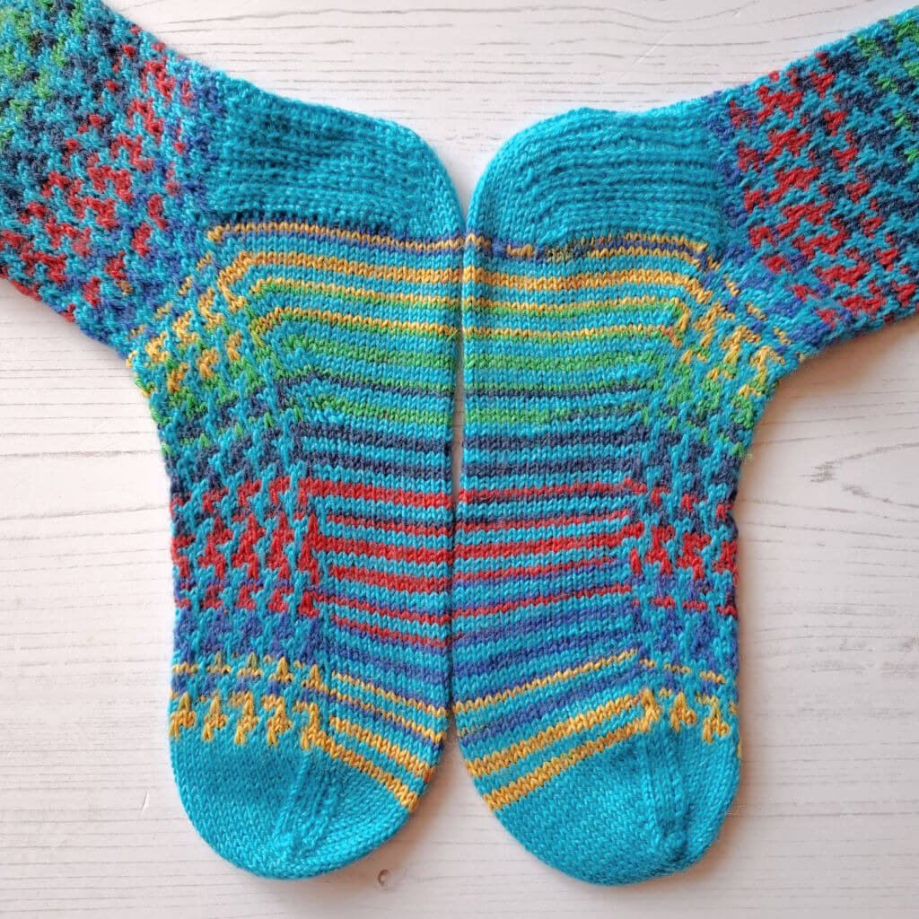 Two colourful knitted socks with intricate patterns rest side by side on a light wooden surface. They feature vibrant blue, red, yellow, and green hues.