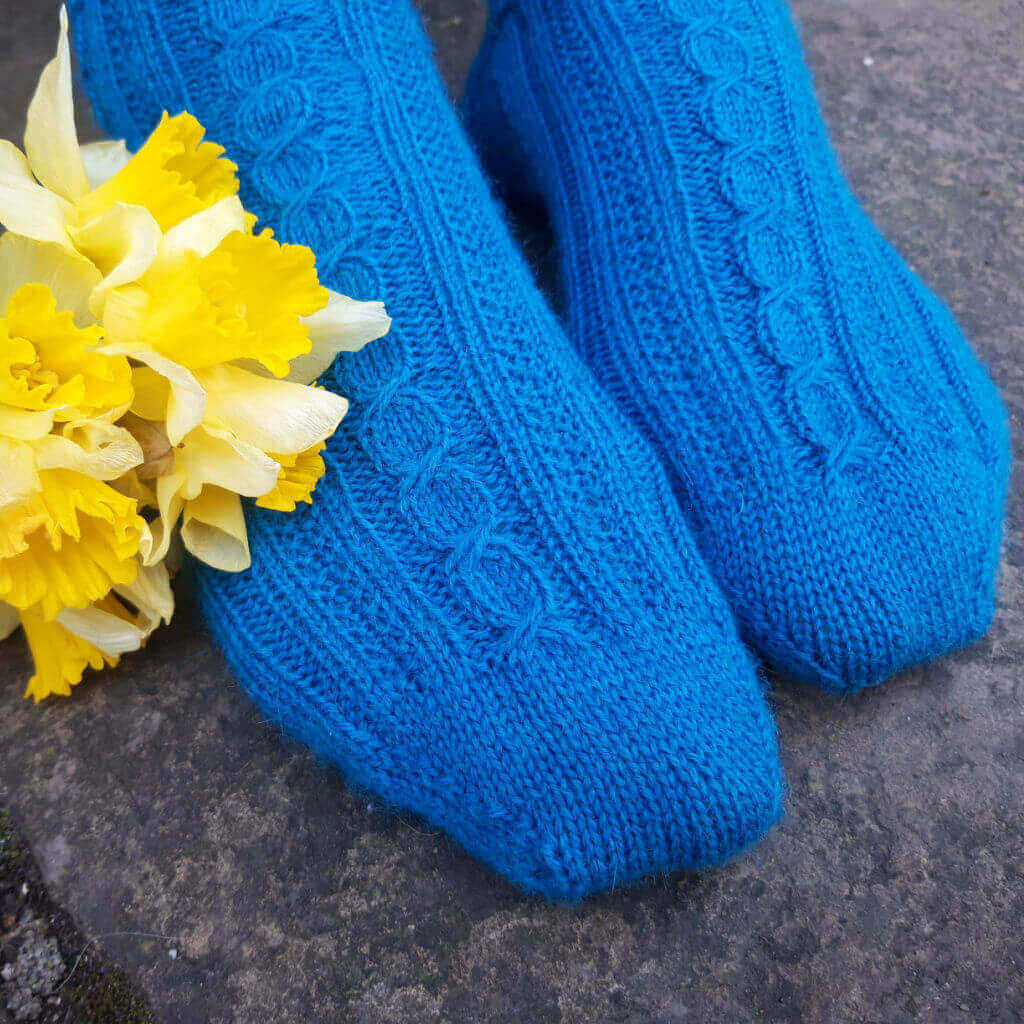 Bright blue knitted socks with intricate patterns are placed on a stone surface. A yellow daffodil bouquet adds a touch of spring vibrancy. Winwick Mum | Amory Socks