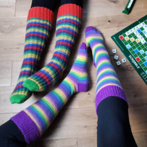 Two pairs of legs in colourful striped socks, one pair rainbow-themed, the other in warm tones, on a wooden floor next to a Scrabble game board. Cosy and playful atmosphere. Winwick Mum | Basic 4ply Knee High Socks | Beginner sock knitting | Free sock knitting patterns