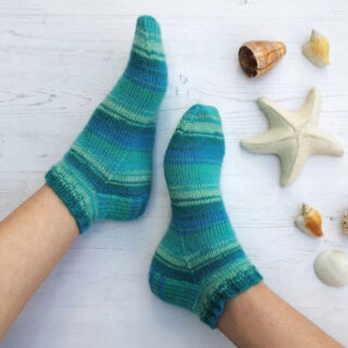 Two feet wearing cosy, teal and blue striped socks rest on a light wooden floor. Nearby are seashells and a starfish, evoking a calm, beachy vibe. Winwick Mum | Basic 4ply Shortie Socks | Beginner sock knitting | Free sock knitting patterns