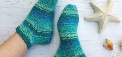 Two feet wearing cosy, teal and blue striped socks rest on a light wooden floor. Nearby are seashells and a starfish, evoking a calm, beachy vibe. Winwick Mum | Basic 4ply Shortie Socks | Beginner sock knitting | Free sock knitting patterns