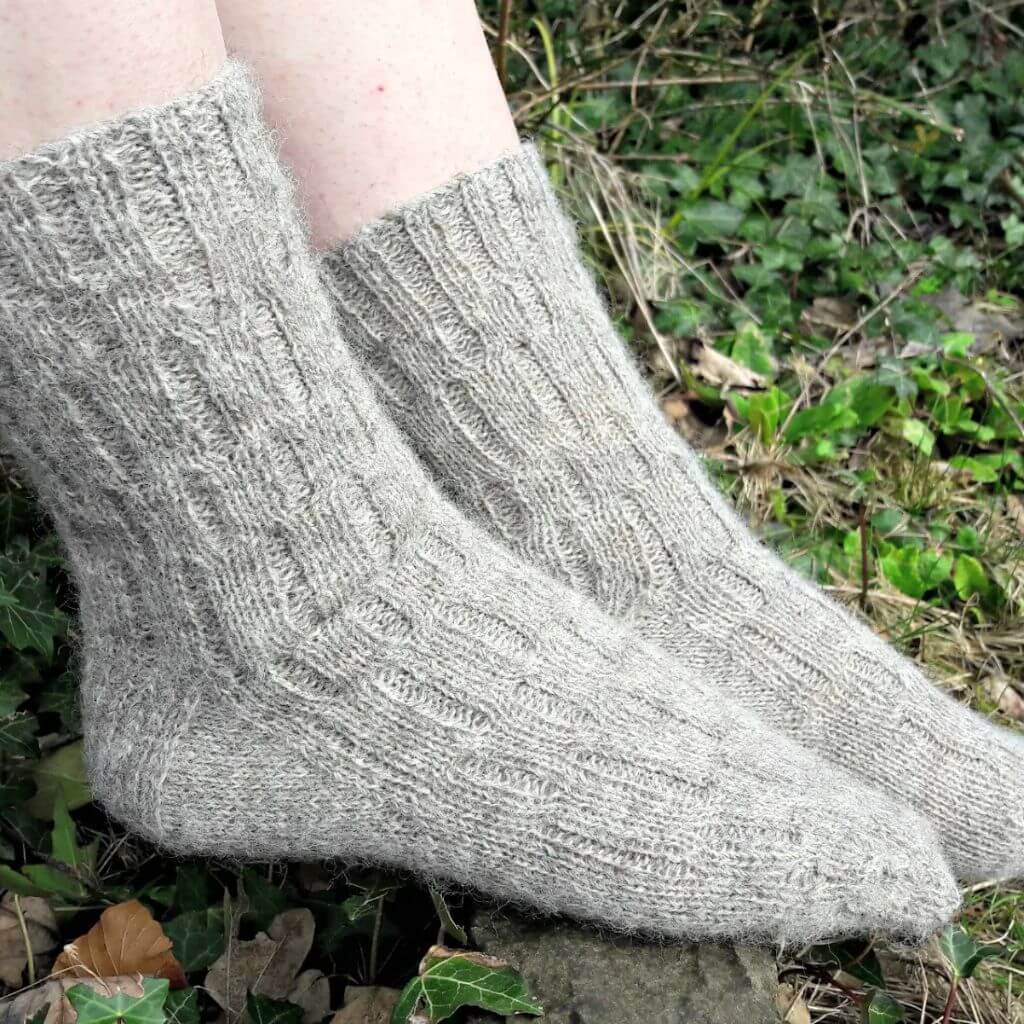 Close-up of two feet wearing thick, textured grey wool socks, resting on mossy ground with scattered green leaves, evoking warmth and coziness. Winwick Mum | Calcite Socks