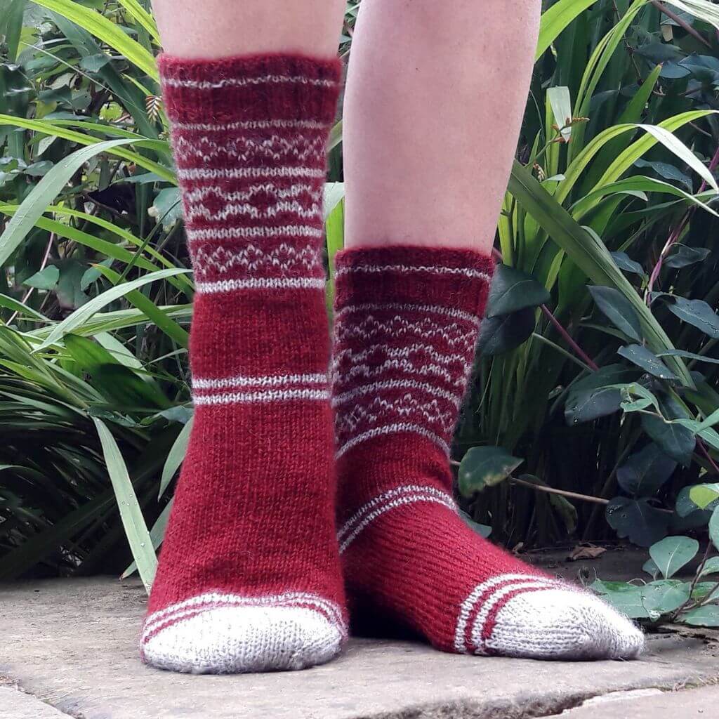 Red knit socks with white geometric patterns and toe detailing on feet, standing on a stone surface. Lush green plants surround in the background. Winwick Mum | Easy Colourwork Socks