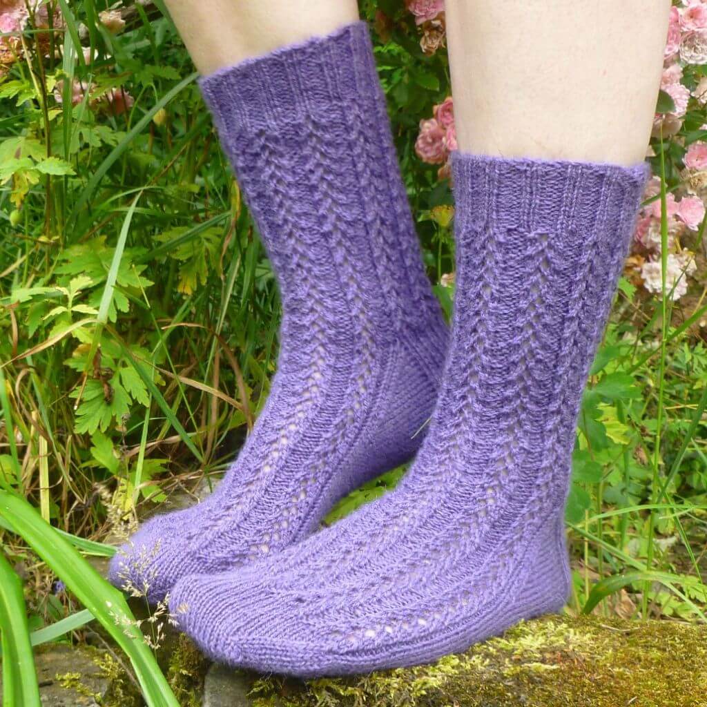 Purple knitted socks with intricate patterns showcased on a pair of feet. The background features lush green foliage and delicate pink flowers. Winwick Mum | Easy Lace Socks