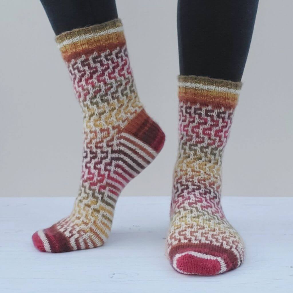 Person wearing colourful knitted socks with a geometric pattern in red, yellow, and brown hues. The background is a neutral, light grey, emphasising the socks. Winwick Mum | Easy Mosaic Socks