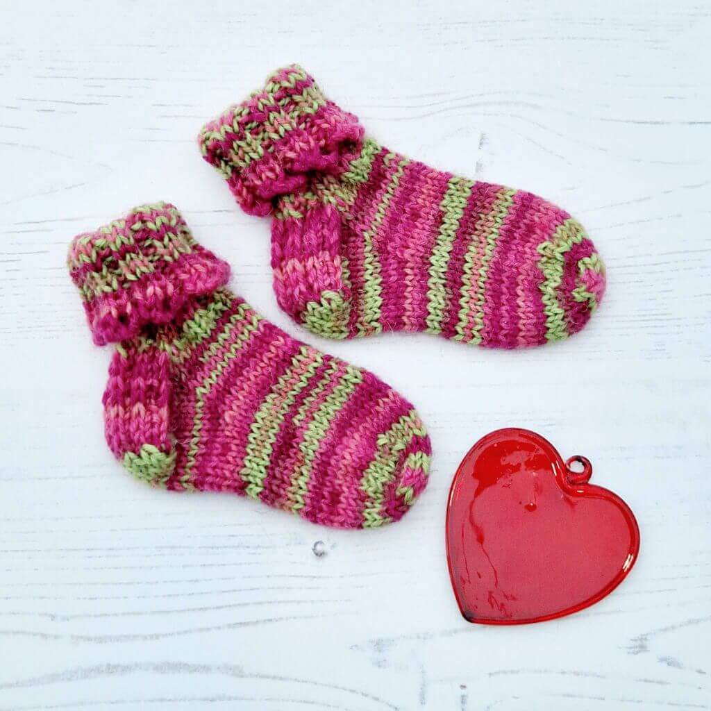 A pair of baby socks in pink and green stripes with a decorative cuff lie on a white background next to a red glass heart. Winwick Mum | Hope Socks