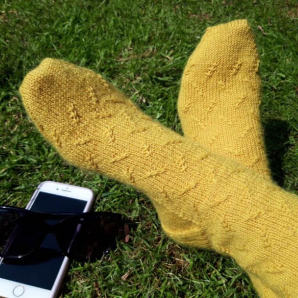 A pair of hand knitted socks in yellow yarn with a twisted stitch pattern. The model is sitting on the grass with feet crossed, and there is a mobile phone and pair of sunglasses next to the feet. Winwick Mum | LikeMinded Socks