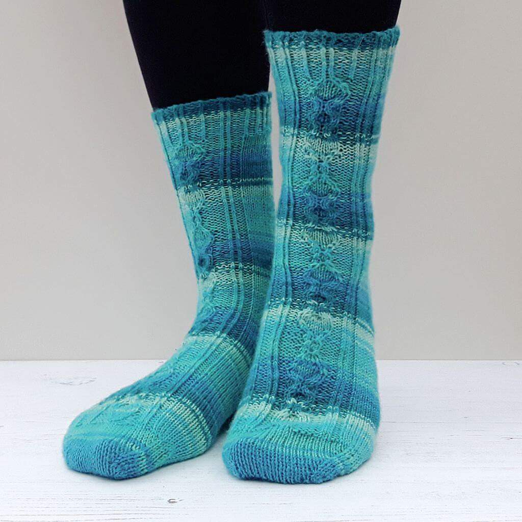 Close-up of feet wearing turquoise and blue blue striped knitted socks with intricate patterns and ribbed cuffs, standing on a white wooden surface, conveying warmth and comfort. Winwick Mum | Love Spoon Socks
