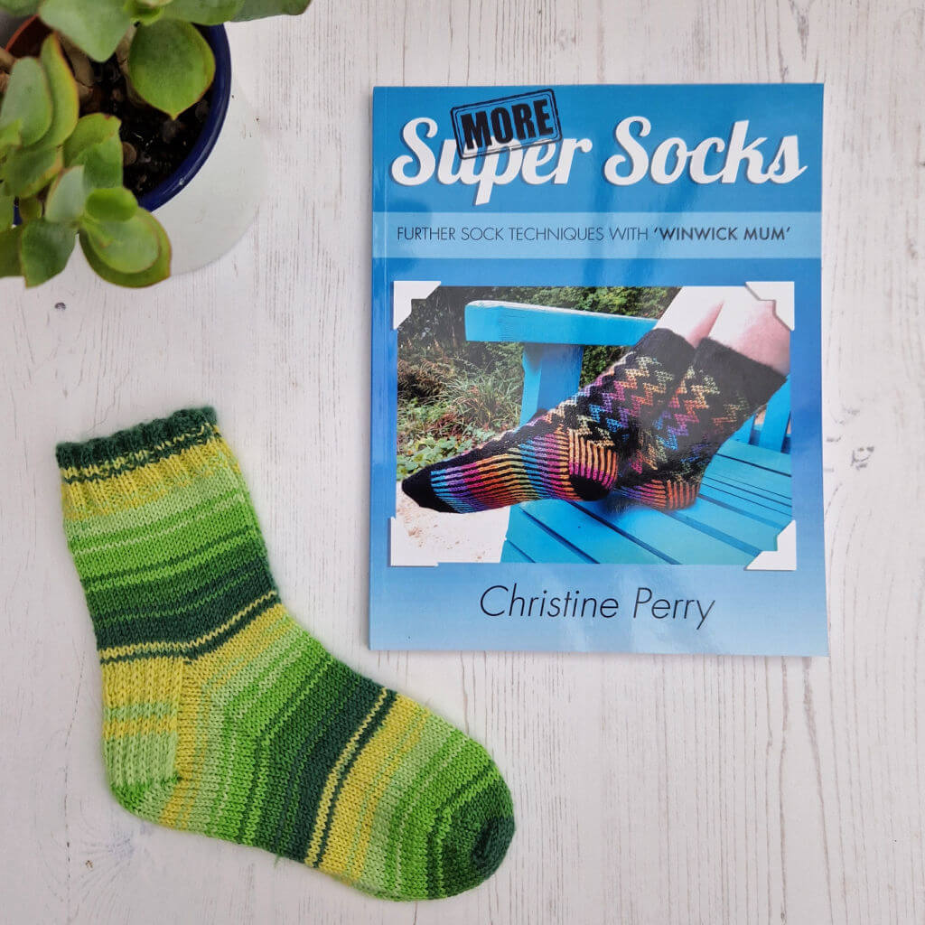 Knitting book titled "More Super Socks" by Christine Perry on a wooden surface, beside a green knitted sock and a potted plant. Bright, crafty scene.