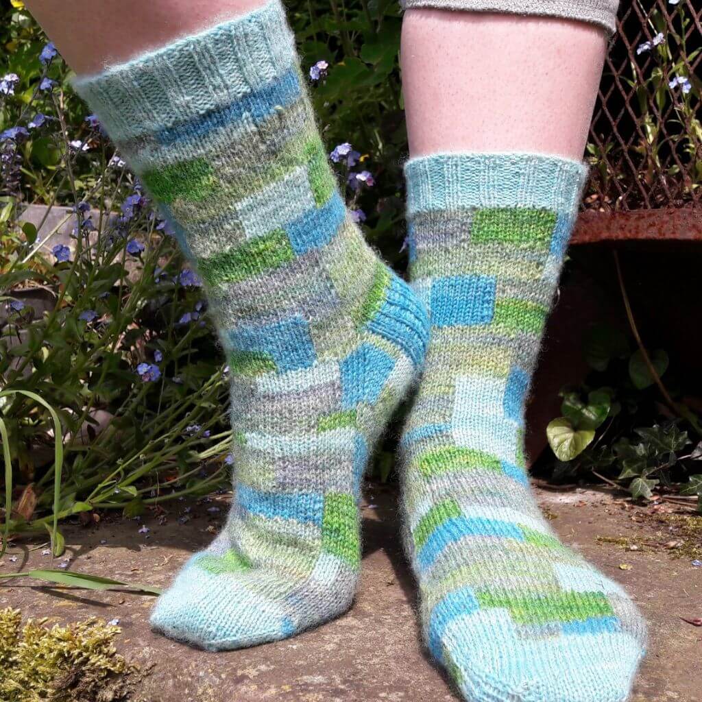 Close-up of legs wearing colourful knitted socks in shades of blue, green, and grey with a patchwork pattern. They stand on a stone path surrounded by small blue flowers and greenery, creating a cozy and serene atmosphere. Winwick Mum | Patchwork Socks