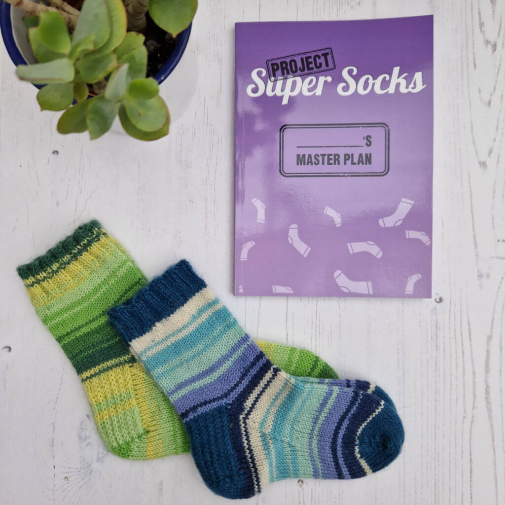 A pair of colorful striped socks lie on a white wooden surface next to a purple book titled "Project Super Socks." A potted plant adds a fresh touch. Winwick Mum | Project Super Socks | sock knitting