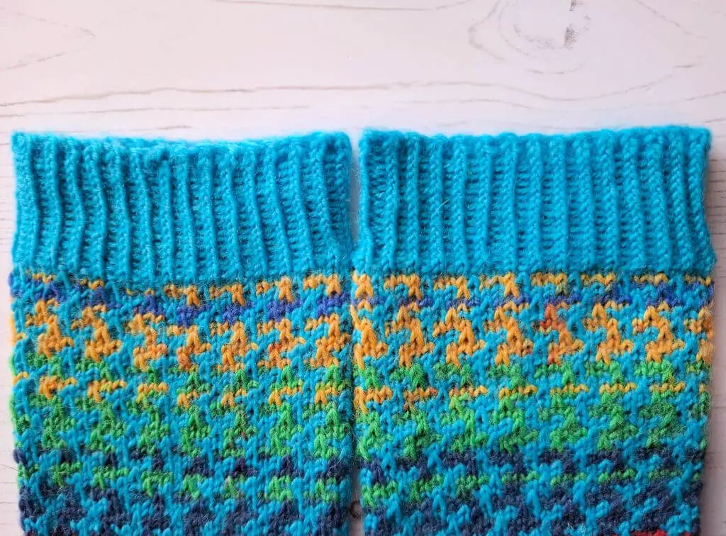 Knitted socks with a ribbed turquoise cuff and colourful mosaic pattern in yellow, green, and blue. Background is a light wooden surface.