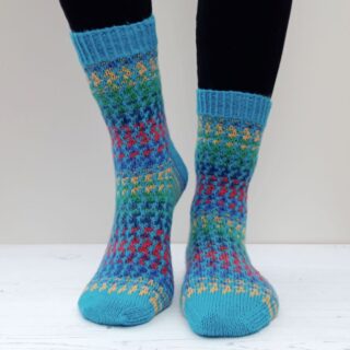 Person wearing colourful knitted socks with intricate patterns of blue, red, green, and yellow, standing on a white wooden surface against a neutral background.